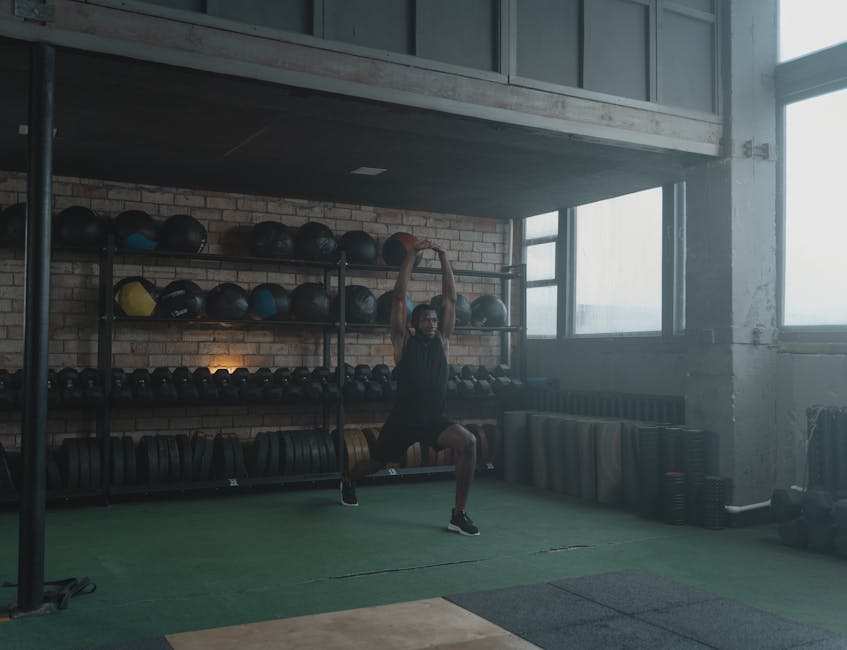 A man performing stretches in a spacious, well-equipped industrial gym environment.